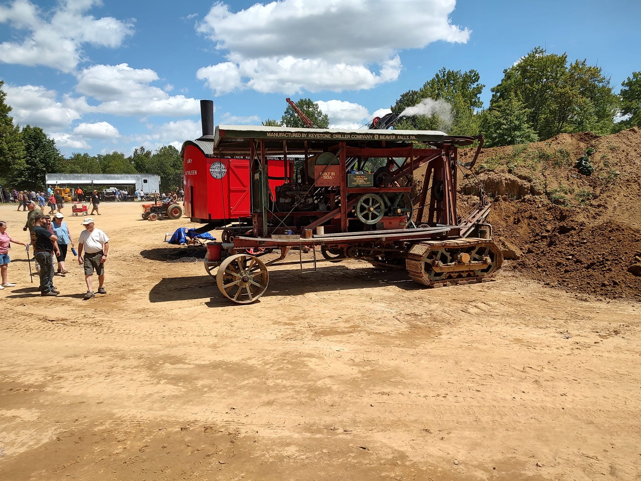 Red steam traction engine on crawler tracks with a steam plume, crowd watching nearby